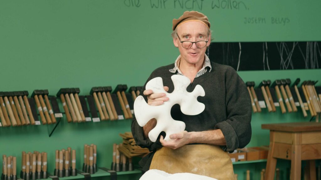 Bildhauer in der Werkstatt von Bildhau KG in Köln. Er trägt einen dunklen Pullover und eine Baskenmütze und präsentiert ein weißes, puzzleteilförmiges Kunstobjekt. An der grünen Wand hinter ihm ist ein Zitat von Joseph Beuys zu sehen.