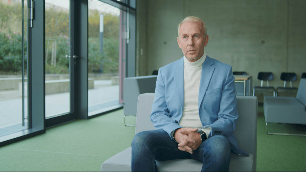 Thomas Höchst, Schulleiter der IGS Contwig, sitzt in der modernen Schulbibliothek. Er trägt einen hellblauen Blazer und weißen Rollkragenpullover und sitzt auf einem grauen Sessel vor bodentiefen Fenstern. Der Raum hat einen mintgrünen Boden und im Hintergrund sind weitere Sitzgelegenheiten zu sehen.