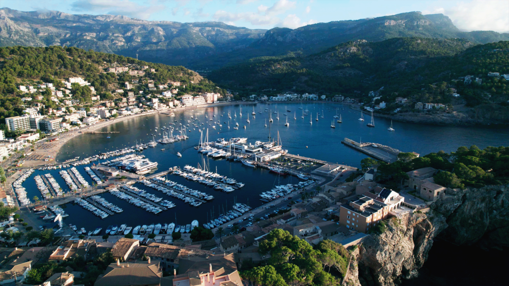 Luftaufnahme des Port de Soller auf Mallorca. Der Yachthafen ist umgeben von bewaldeten Bergen und terrassenförmig angelegten Häusern. Zahlreiche weiße Segelboote und Yachten liegen in den geometrisch angeordneten Anlegestellen. Das türkisblaue Wasser kontrastiert mit der üppigen grünen Vegetation.
