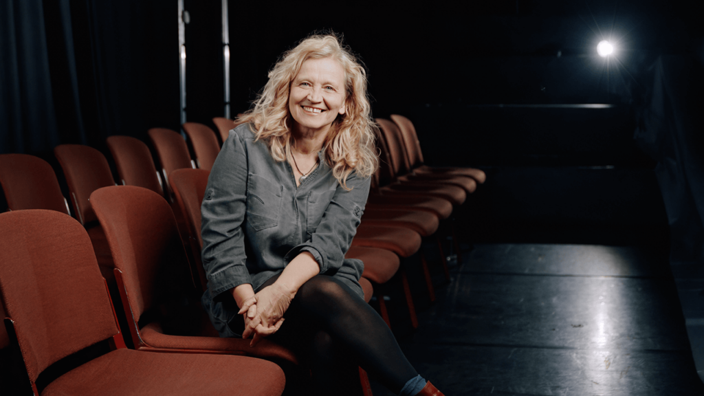 Bettina Kaminski, Leiterin des Freien Schauspiel Ensembles Frankfurt, sitzt lächelnd in den Zuschauerrängen ihres Theaters. Sie trägt graue Kleidung, im Hintergrund sind die roten Theatersitze und ein Bühnenscheinwerfer zu sehen.
